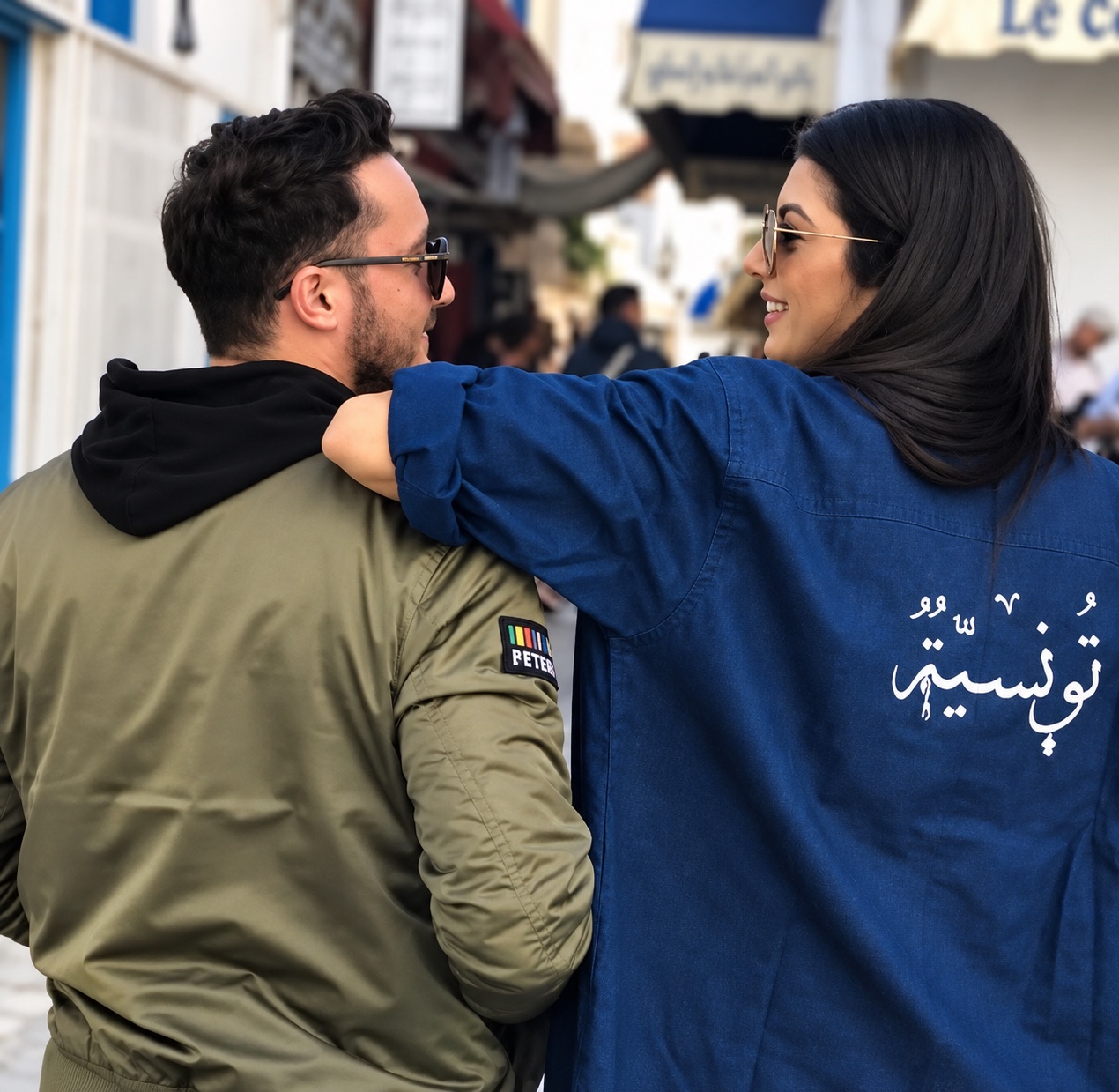 A young couple walking arm in arm through a Tunisian street; her jacket reads Tounsya in Arabic